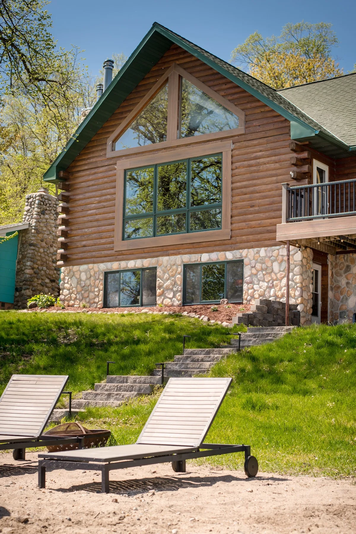 Cabin exterior from the beach showing A-frame log construction, stone foundation, lounge chairs, and fire pit