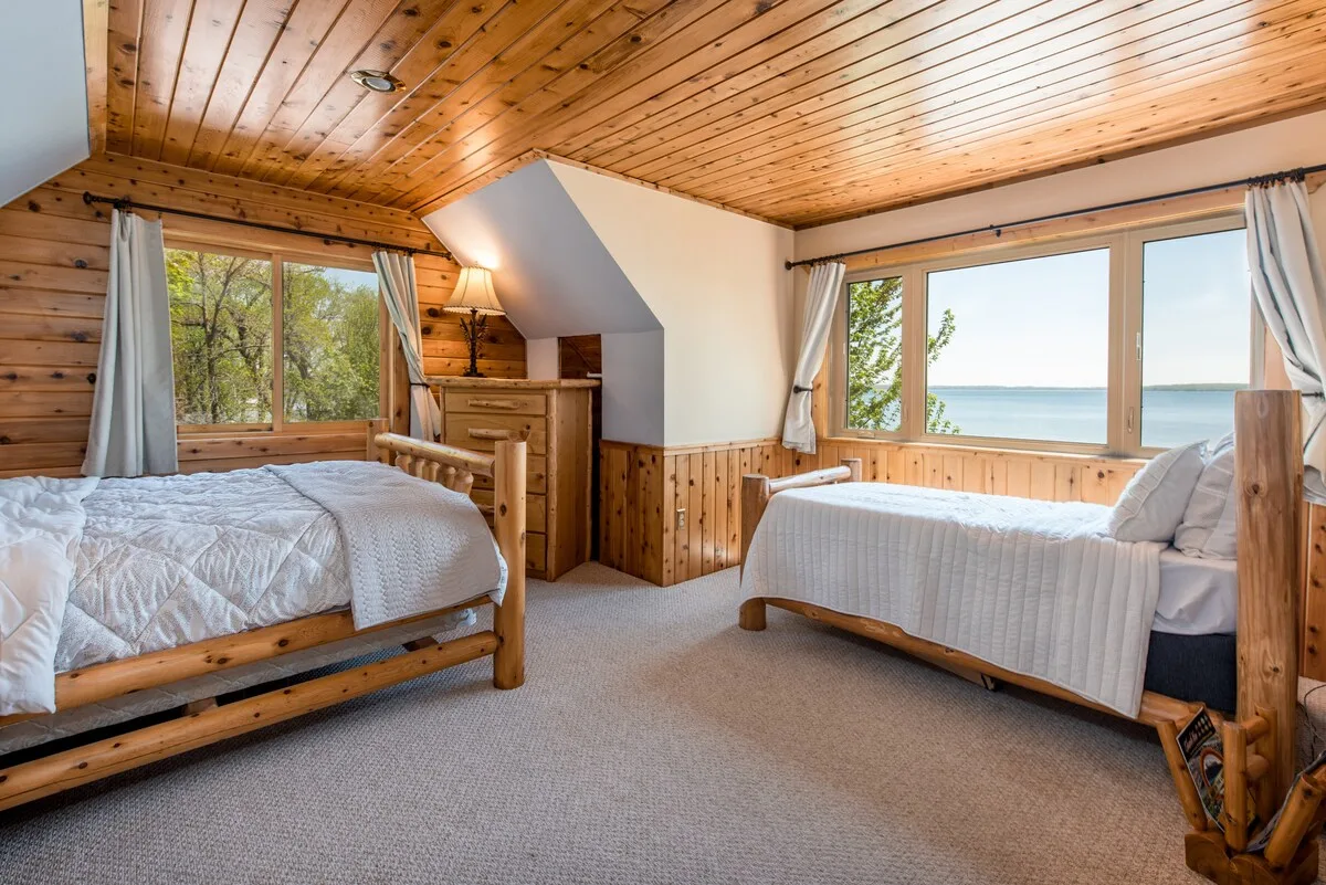Sandbar Suite with two log-frame beds, cedar ceiling, and panoramic lake views from both windows