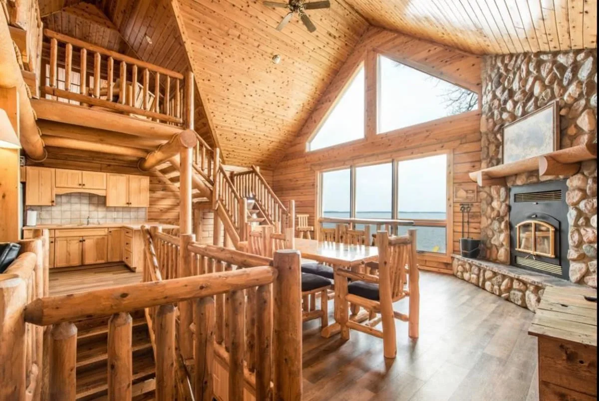 Open-concept great room with soaring log ceilings, loft balcony, stone fireplace, and panoramic Lake Miltona views